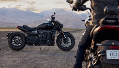 A Triumph Rocket 3 R Storm motorcycle parked on an open road with mountainous landscape in the background, showcasing its robust design and black finish.