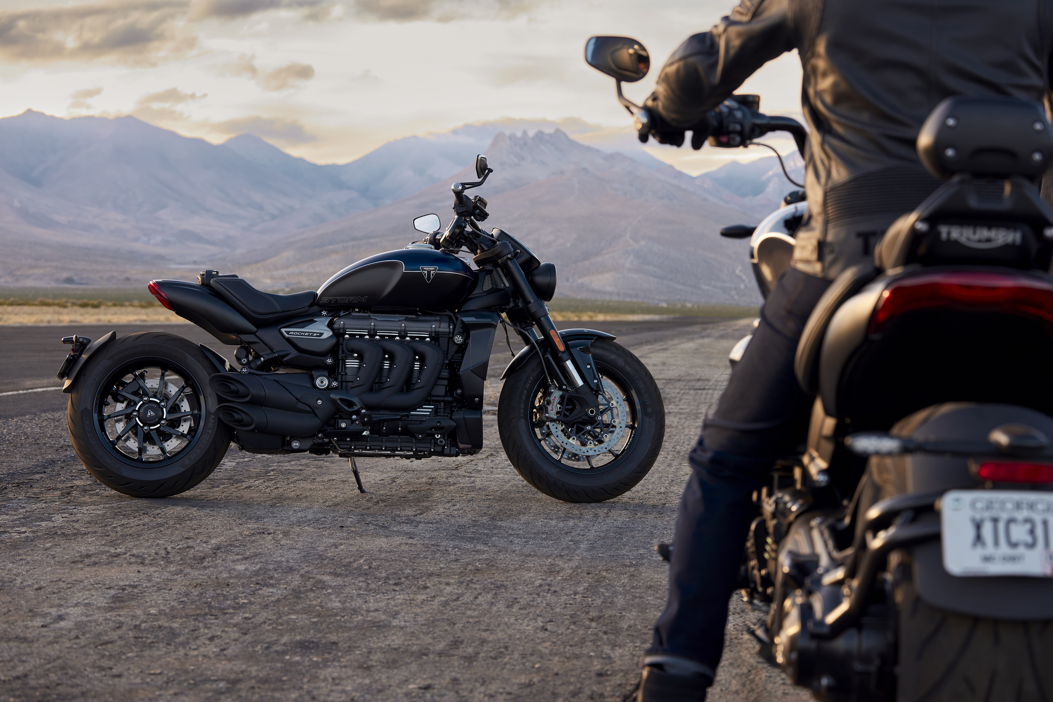 A Triumph Rocket 3 R Storm motorcycle parked on an open road with mountainous landscape in the background, showcasing its robust design and black finish.